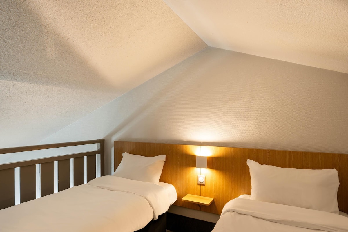Une chambre d’hôtel avec deux lits, une table de chevet, une lampe fixée au mur et un plafond en pente.