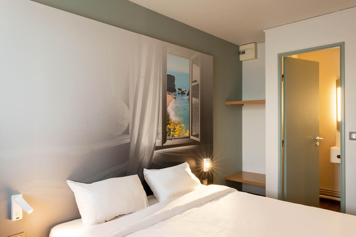 Une chambre d’hôtel avec un lit, une table de chevet, une lampe fixée au mur, une fenêtre donnant sur la mer et une porte.