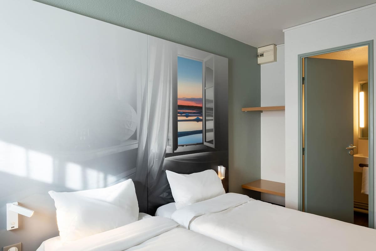 Une chambre d’hôtel comprenant un lit, une table de chevet, une lampe fixée au mur et une porte donnant accès à une salle de bain.