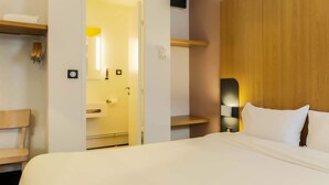 Una habitación de hotel con una cama, cabecera de madera, estante de madera, espejo, toallero y una puerta que da a un baño.