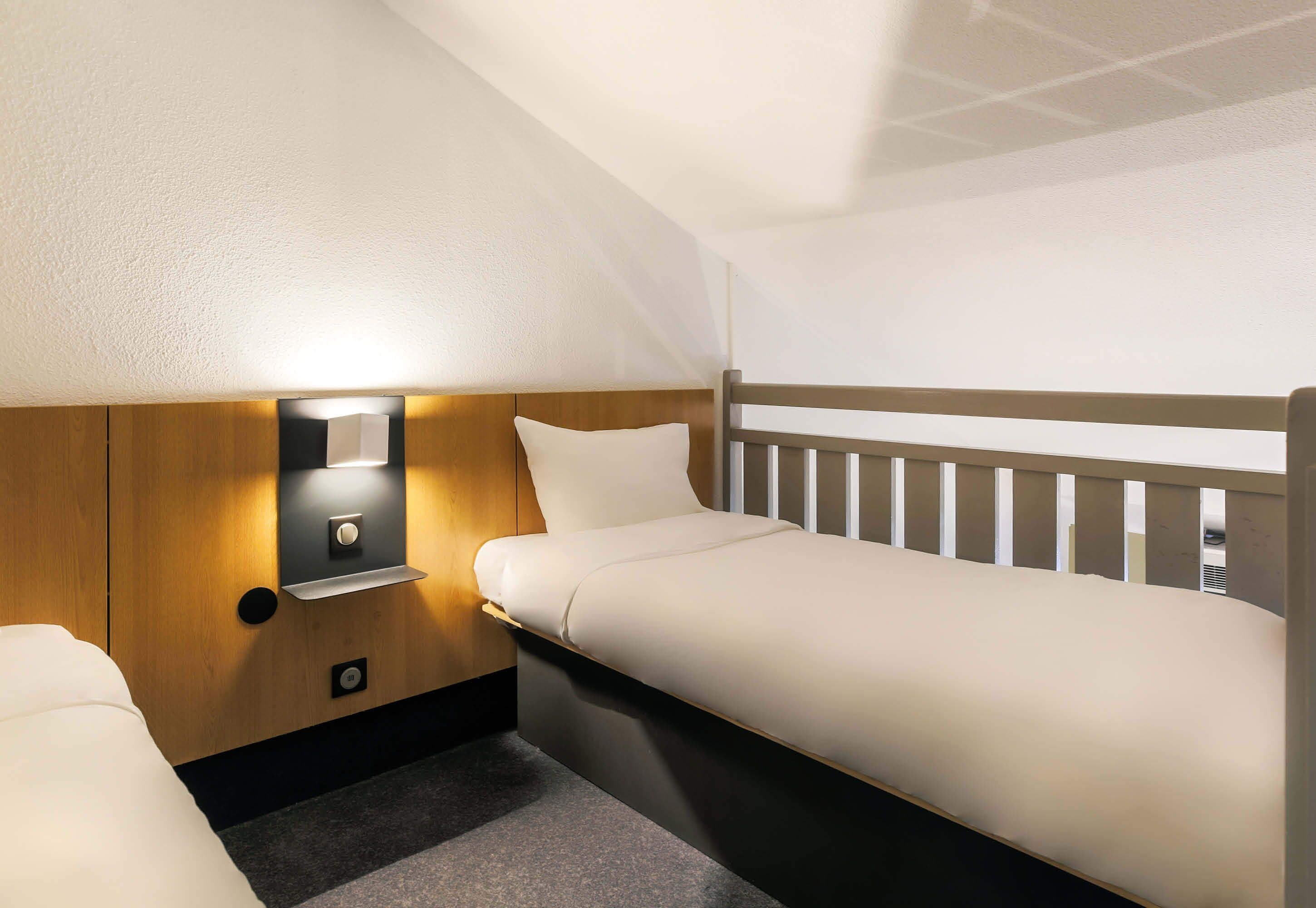 A compact hotel room with a single bed, a nightstand, and a wall-mounted light.