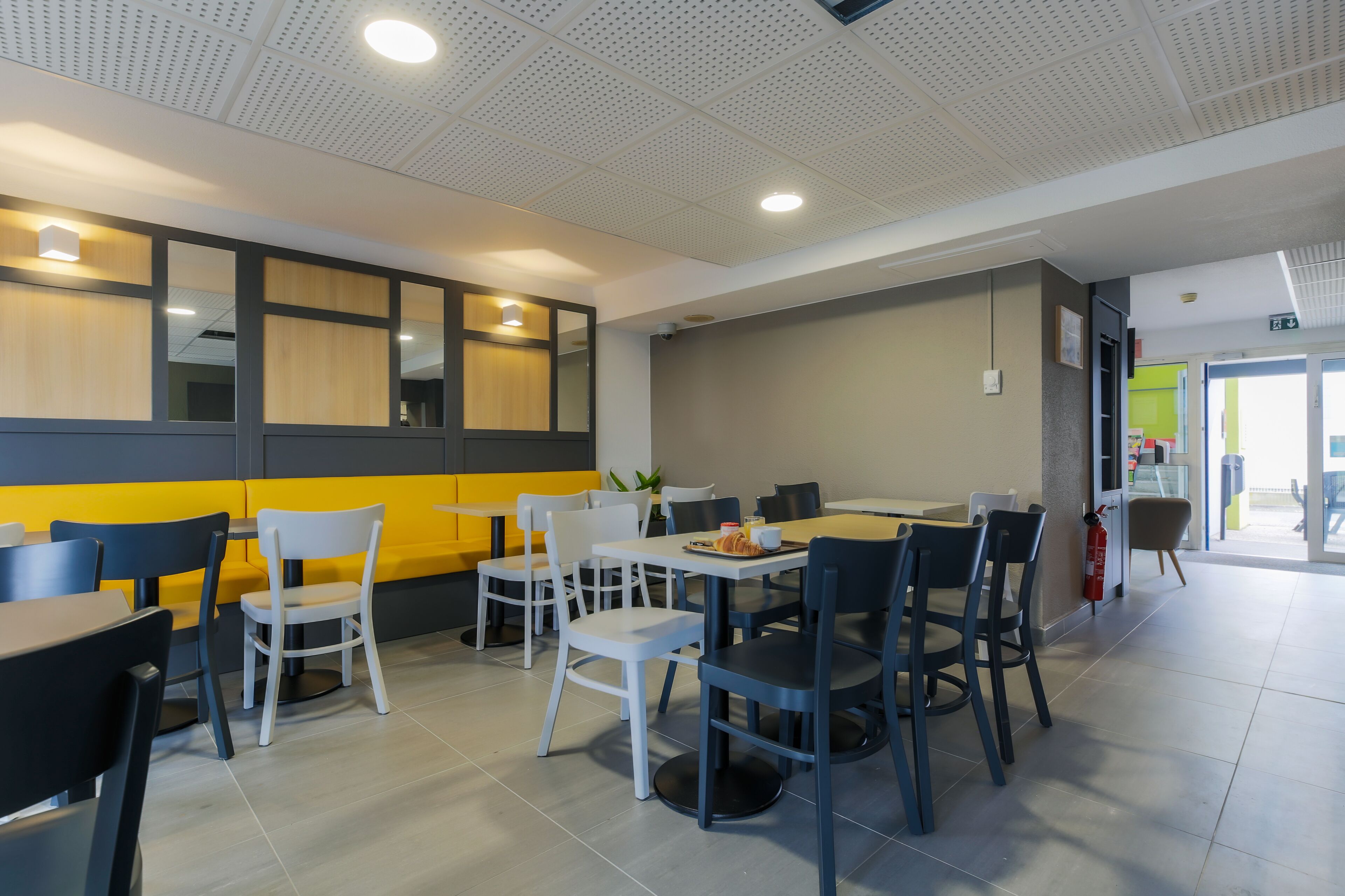 A modern restaurant with a mix of white and dark chairs, yellow and grey walls, and a tiled floor.