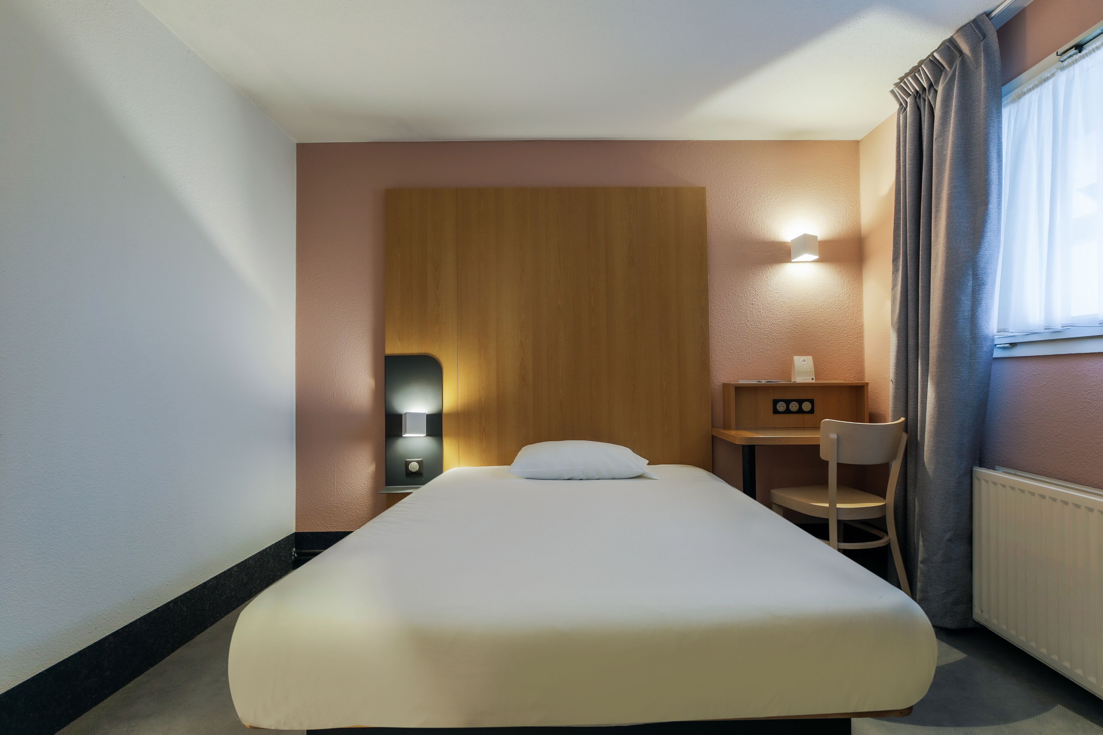 Habitación de hotel con cama, escritorio y silla, luz en la pared y ventana con cortinas.