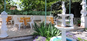Un patio al aire libre con sillas y mesas de plástico blanco, una fuente blanca y una valla verde al fondo.