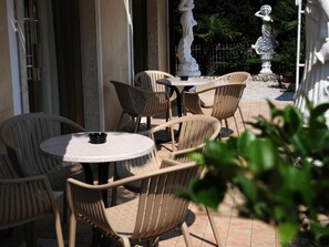 Een terras buiten met ronde tafels en rieten stoelen, omgeven door een tuin met beelden en een hek.