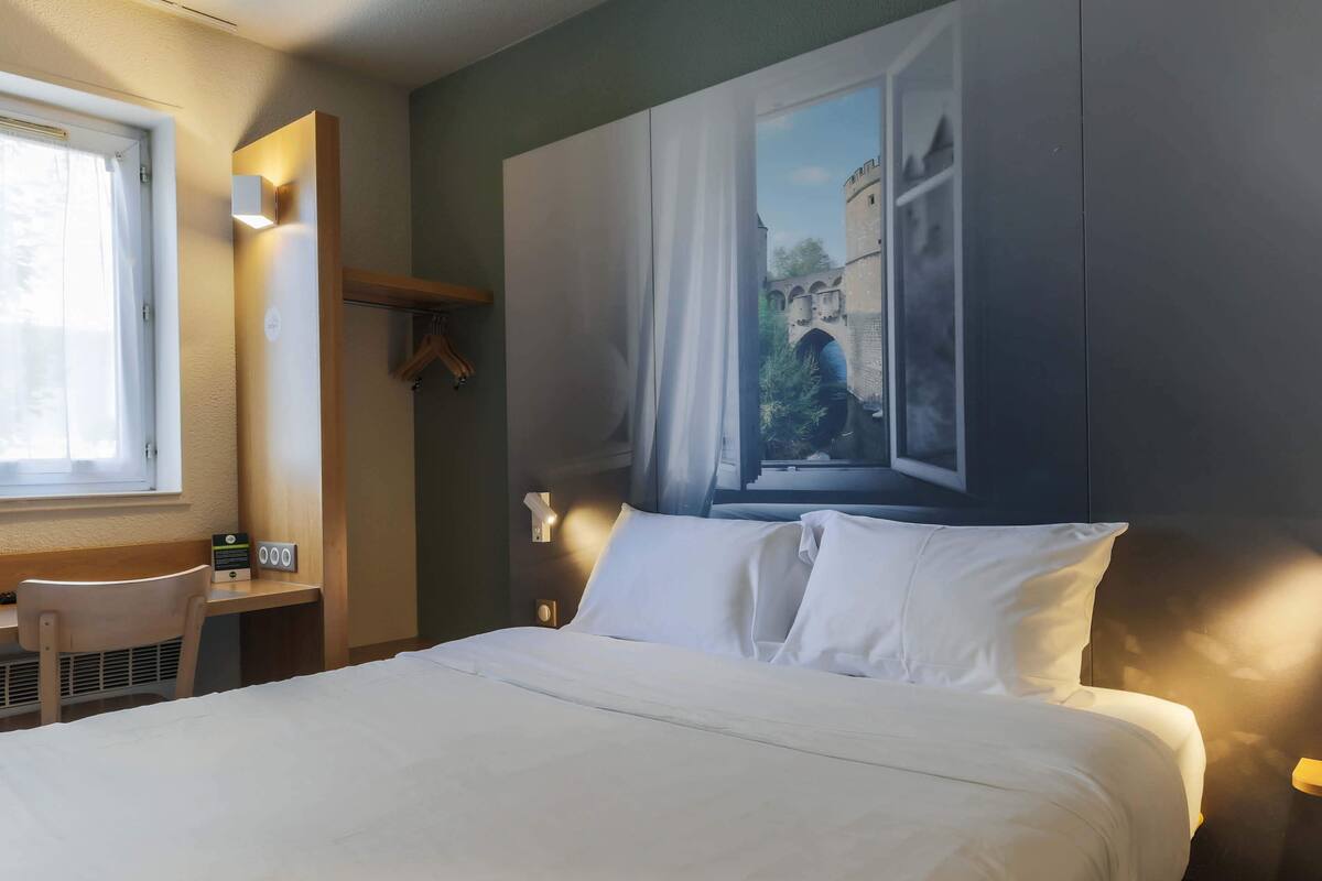 Ein Hotelzimmer mit Bett, Schreibtisch mit Stuhl, Fenster mit Vorhängen und einer Wandlampe.