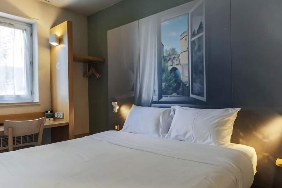 Quarto de hotel com cama, escrivaninha, cadeira, janela e um quadro na parede.