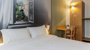 A hotel room with a bed, a desk, and a view of a castle through a window. - B&B HOTEL Metz Jouy-aux-Arches (Jouy-aux-Arches)
