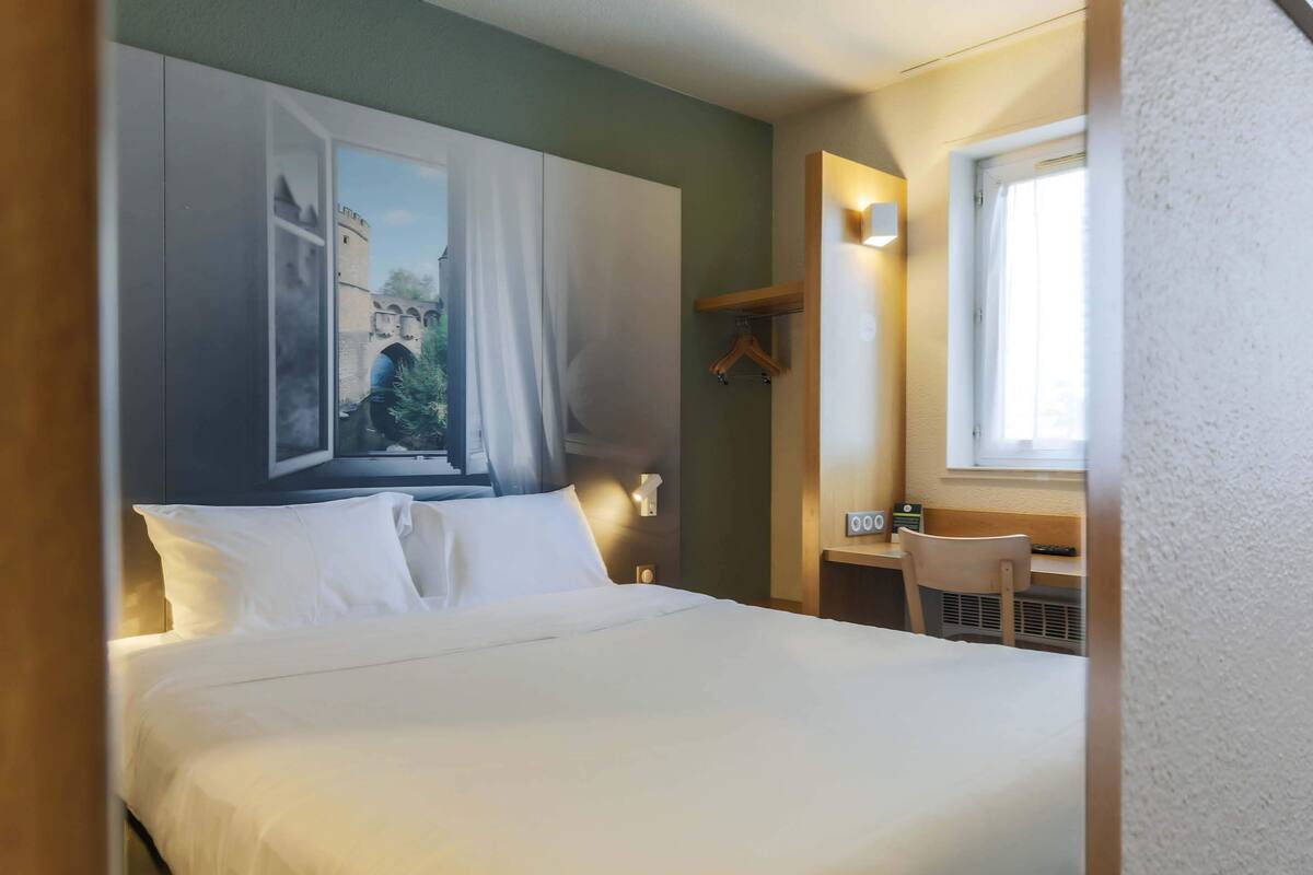 Ein Hotelzimmer mit einem Bett, einem Schreibtisch, einem Sessel, einem Fenster mit Vorhängen, einer Wandleuchte und einem Gemälde eines Schlosses.