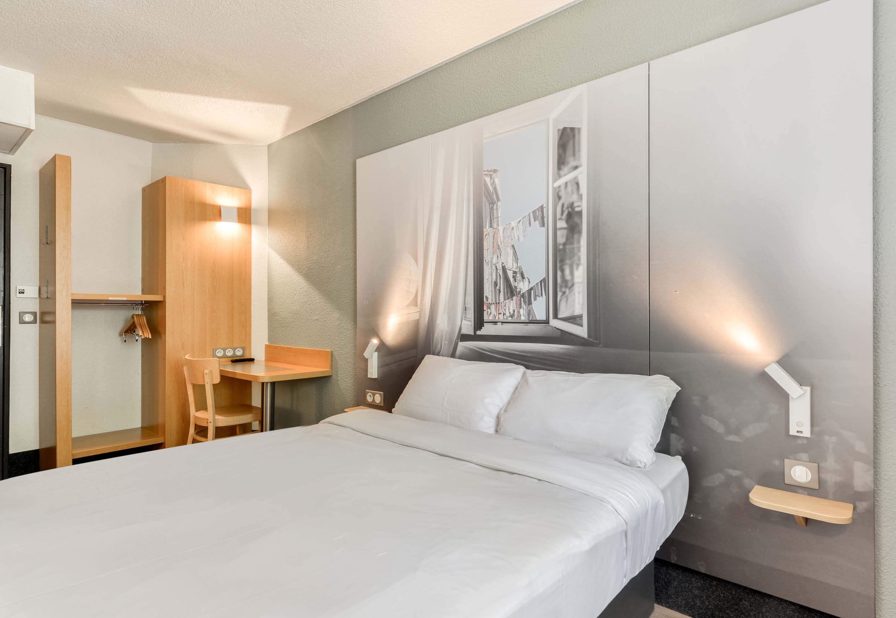 A modern hotel room with a bed, bedside table, chair, and a wall-mounted light fixture.