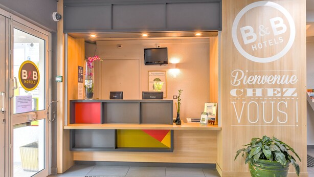 A reception area with a wooden reception desk, a television, and a welcome sign. - B&B HOTEL Montpellier (1) (Saint-Jean-de-Vedas)