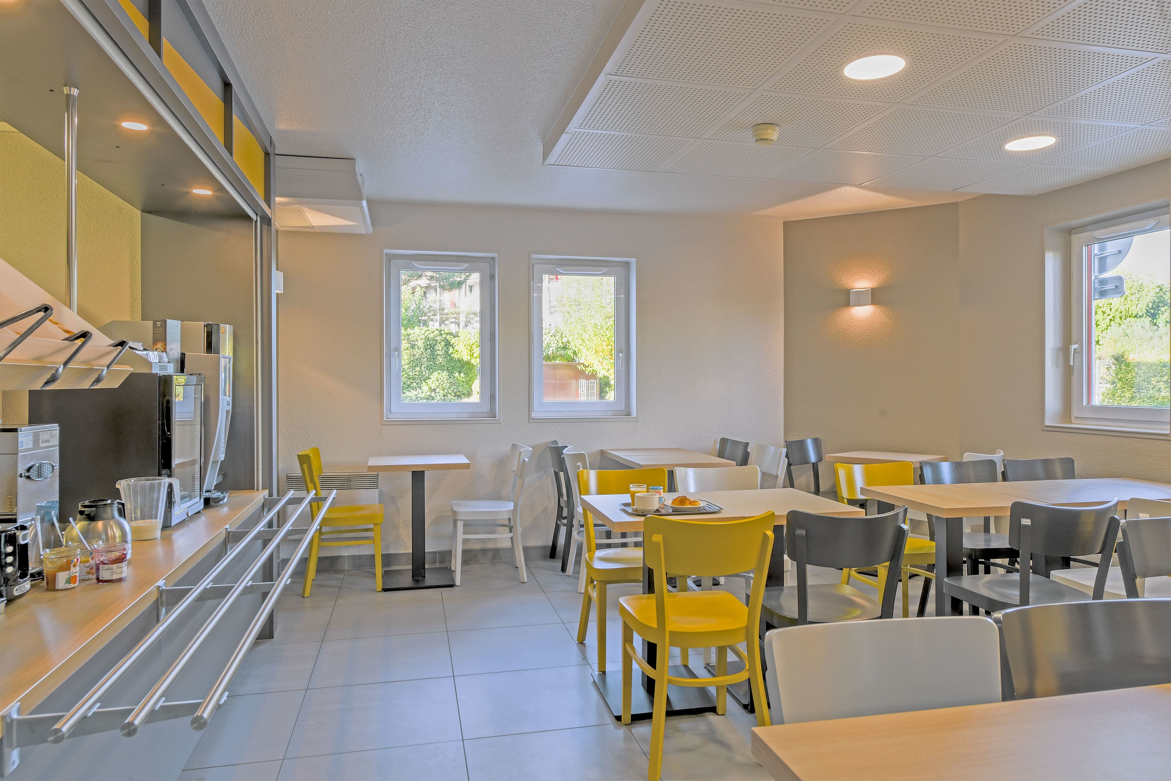 Un comedor moderno con mesas y sillas, una cocina con electrodomésticos, y ventanas que ofrecen luz natural.