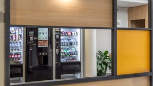 A vending machine area with multiple beverage dispensers, a small plant, and a yellow partition. - B&B HOTEL Montpellier (1) (Saint-Jean-de-Vedas)