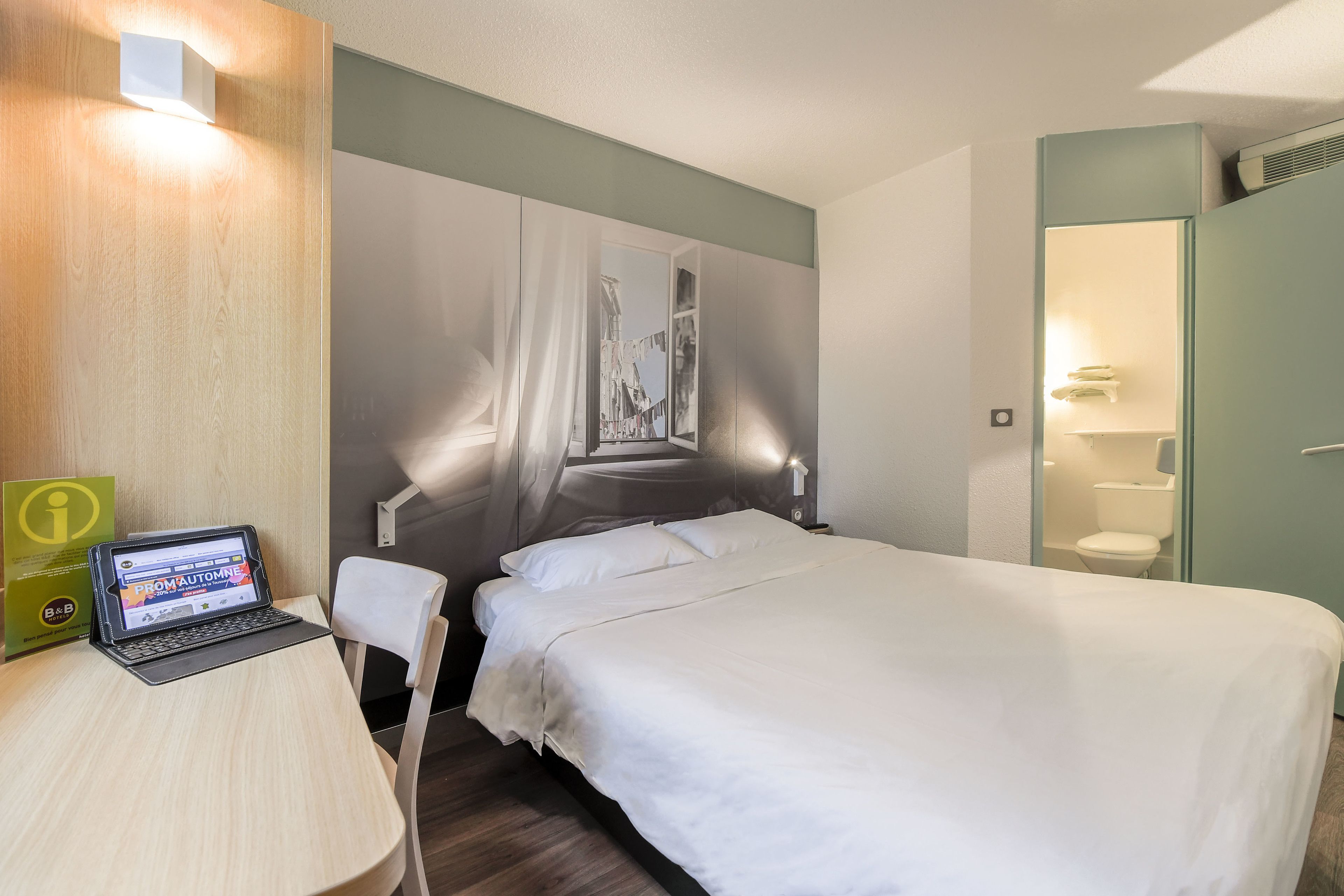 Une chambre d’hôtel comprenant un lit, un bureau avec un ordinateur portable, une salle de bain avec des toilettes et un lavabo, ainsi qu’une grande fenêtre donnant sur un immeuble.