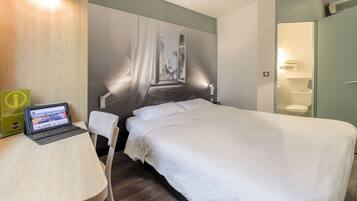 Une chambre d’hôtel comprenant un lit, un bureau avec un ordinateur portable, une salle de bain avec des toilettes et un lavabo, ainsi qu’une grande fenêtre donnant sur un immeuble.