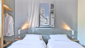 A hotel room with two beds, a view of a historic building through an open window, and a wardrobe with hanging clothes. - B&B HOTEL Montpellier (1) (Saint-Jean-de-Vedas)