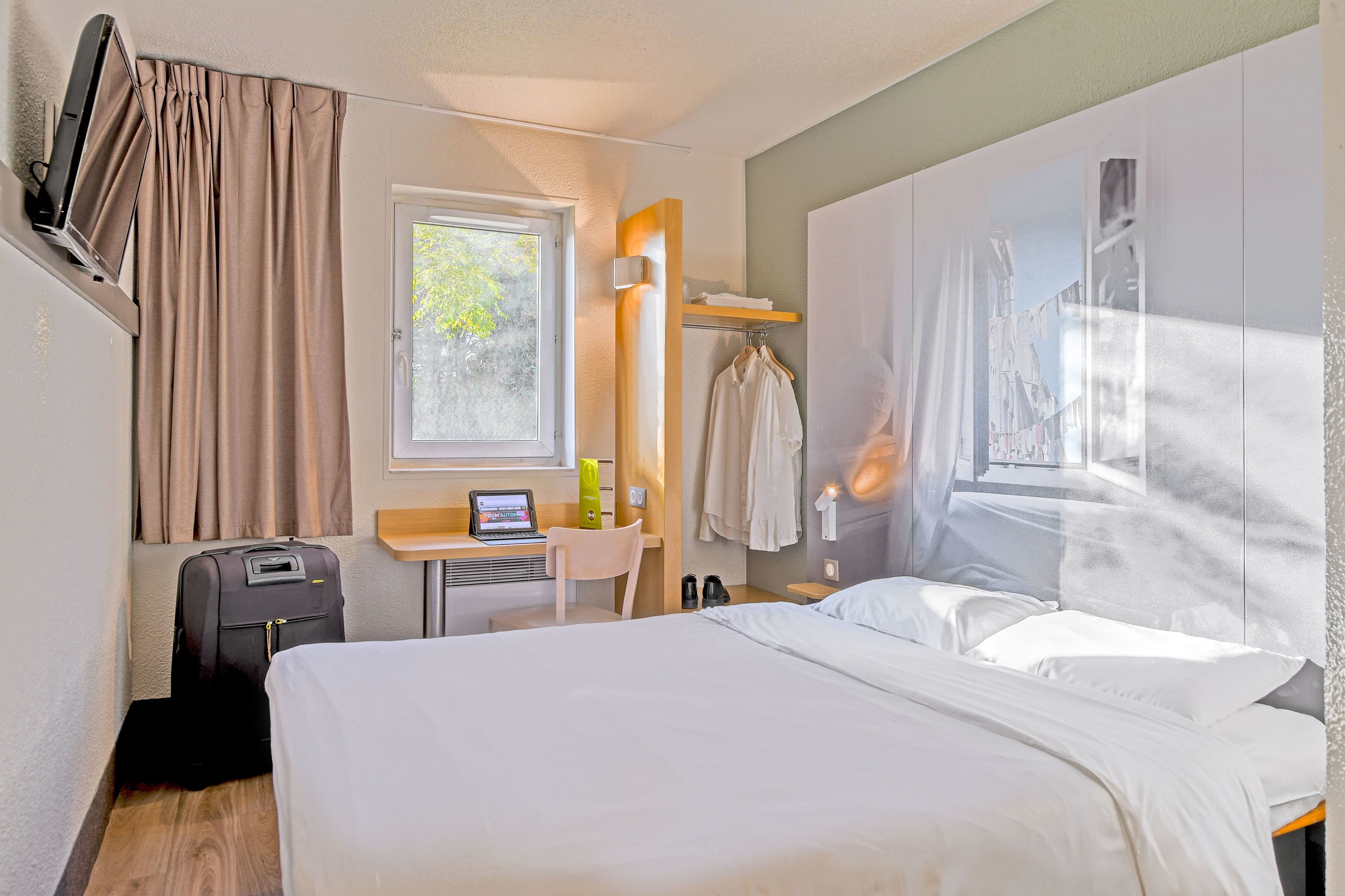 Ein Hotelzimmer mit Bett, Schreibtisch mit Laptop, Koffer, Fenster mit Vorhängen und einem an der Wand befestigten Fernseher.