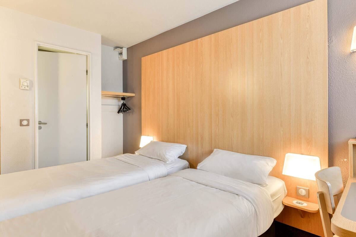 Een hotelkamer met twee bedden, een houten hoofdbord, nachtlampjes, een klein tafeltje en een stoel.