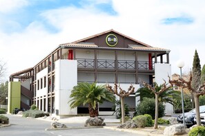 A B&B hotel with a modern design, featuring a prominent B&B logo on the facade, a covered outdoor seating area, and a parking lot with several cars. - B&B HOTEL Montpellier (2) (Saint-Jean-de-Vedas)