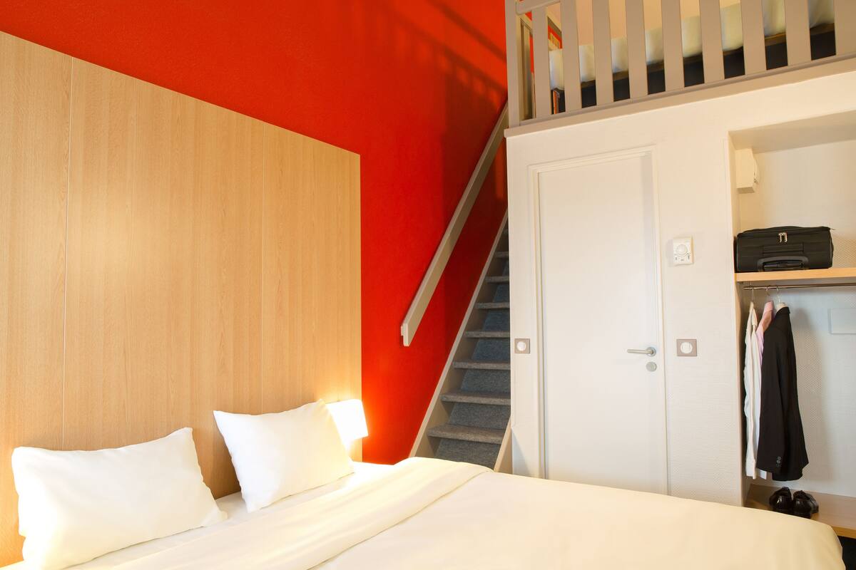 Hotelkamer met een bed, een houten hoofdbord, een rode accentmuur, een trap en een garderobe met kleding.