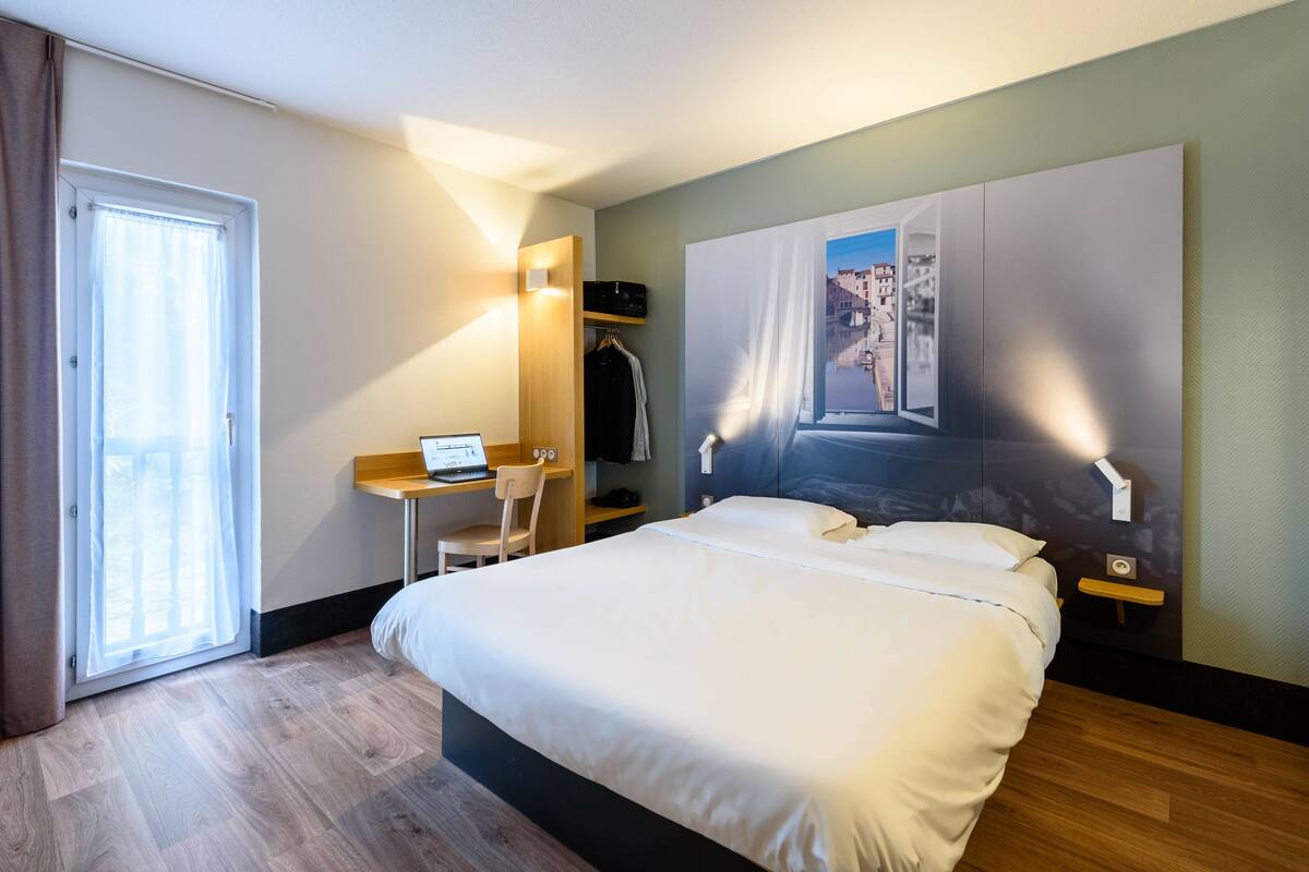 Une chambre d’hôtel avec un lit, un bureau avec un ordinateur portable, une chaise, une armoire et une grande fenêtre.