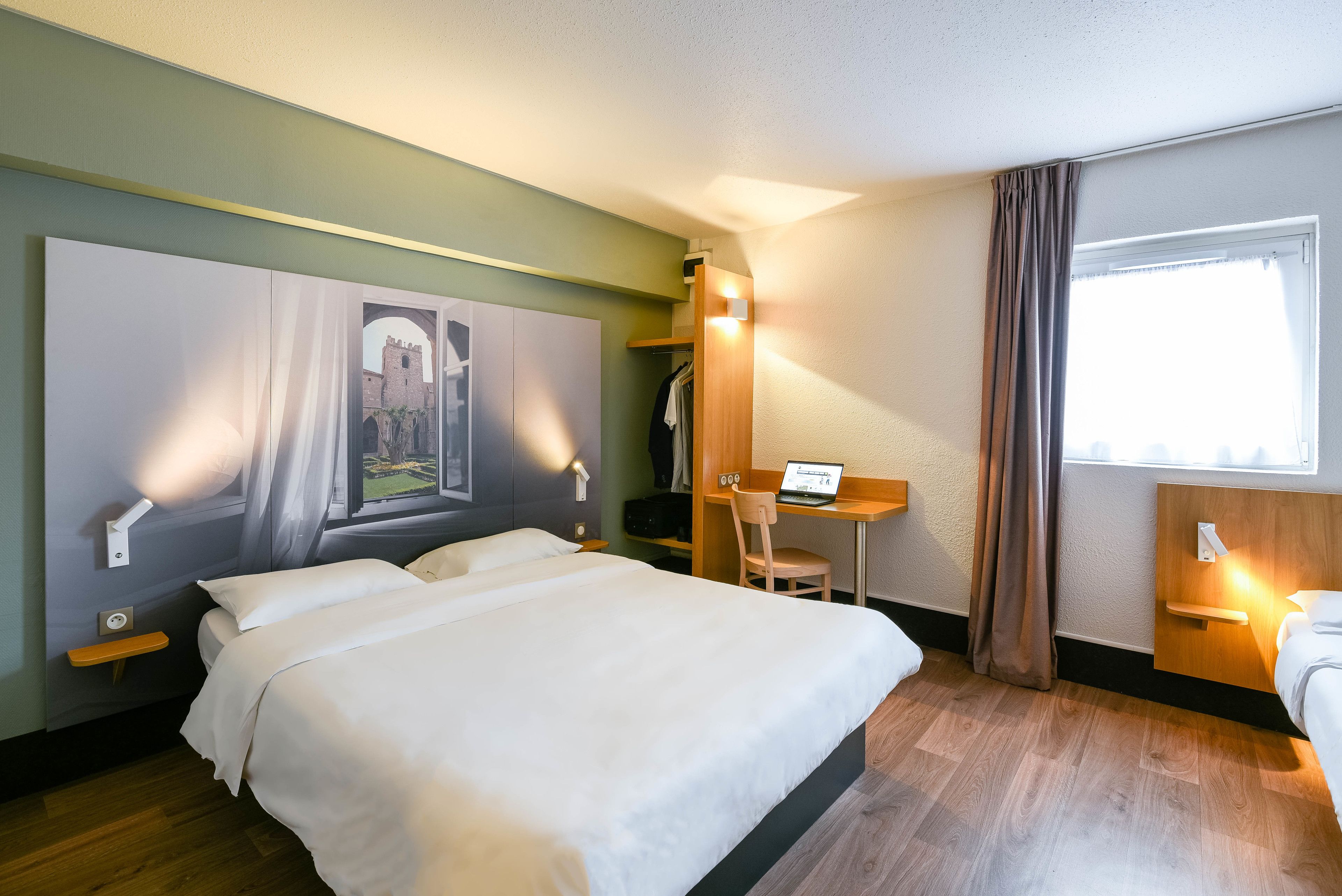 A hotel room with a large bed, a desk, and a chair. There is a painting on the wall and a window with curtains.