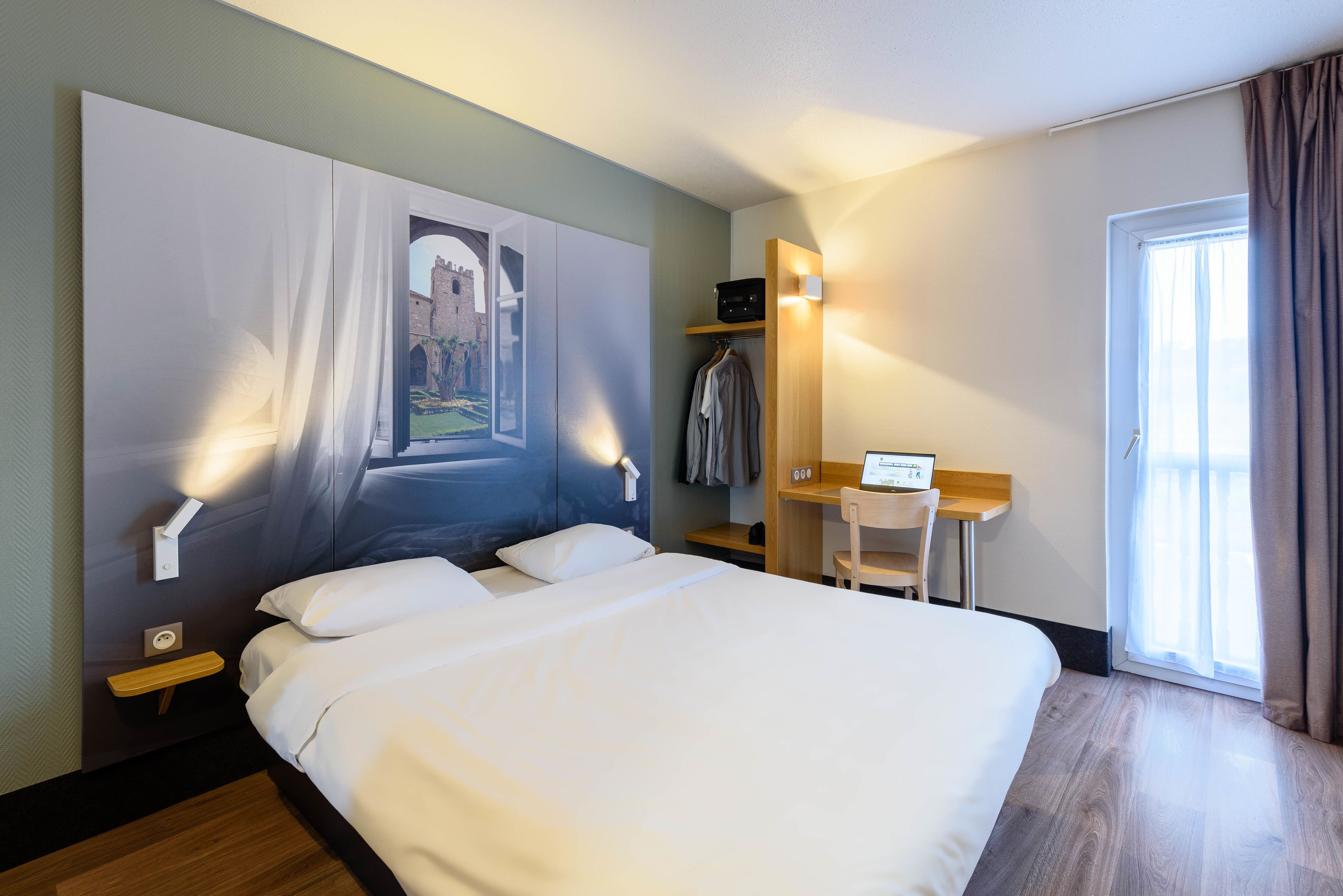 A hotel room with a large bed, a desk, a chair, a coat rack, and a painting on the wall.