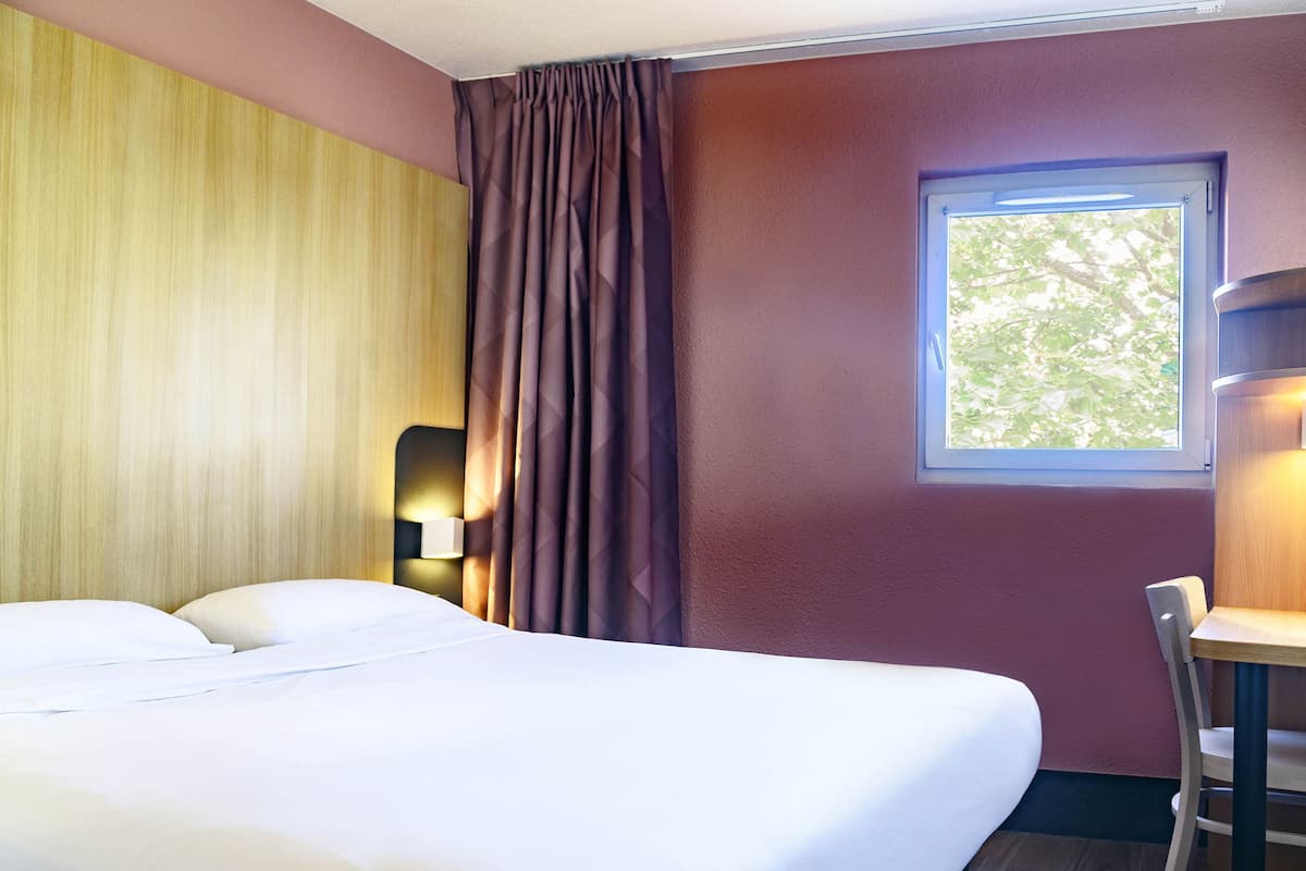 A hotel room with a bed, a desk, and a window with a view of trees.