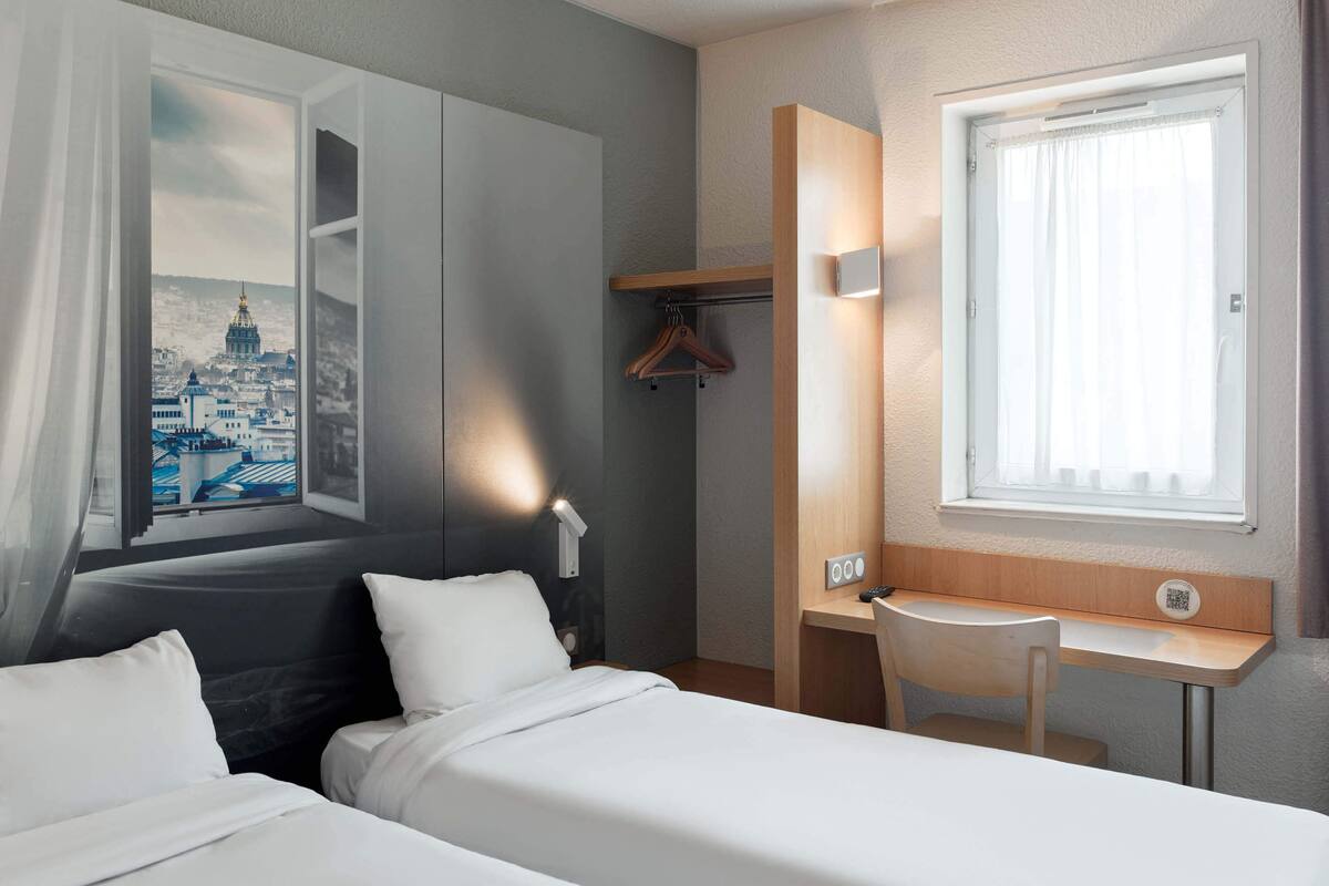 A hotel room with two beds, a desk, a chair, a coat rack, and a window with curtains.