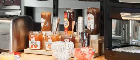 A breakfast bar with various food items and beverages, including jars of jam, a basket of bread, and a coffee machine.