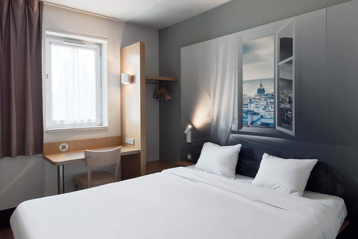 A hotel room with a bed, a desk, a chair, a window with curtains, and a wall-mounted light.
