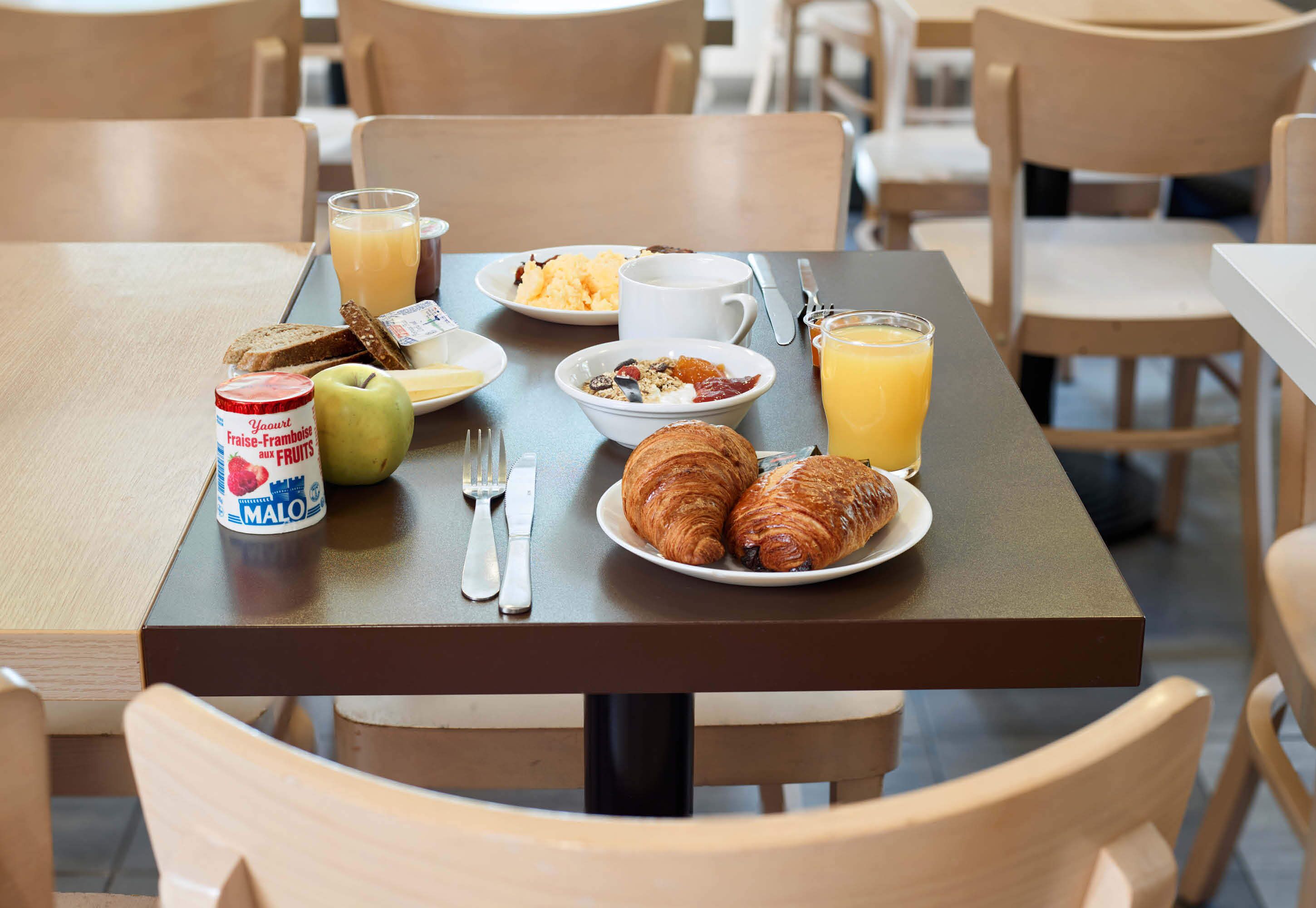 A breakfast table with croissants, a bowl of cereal, a glass of orange juice, and a jar of yogurt.