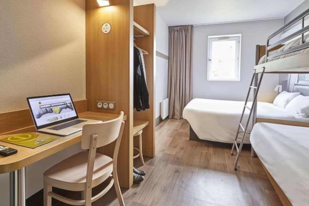 Una habitación de hotel con literas, un escritorio con una computadora portátil, una silla, un armario y una ventana con cortinas.