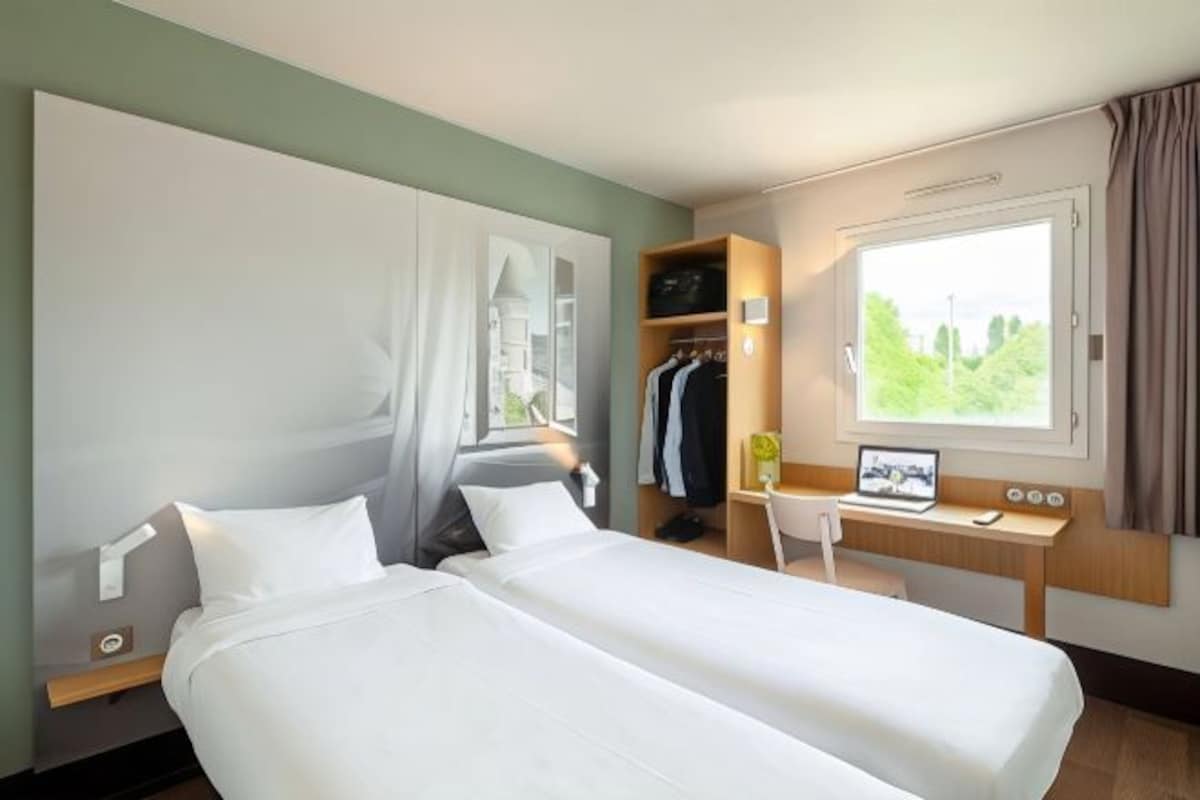 Habitación de hotel con dos camas, un escritorio con una computadora portátil, un armario y una ventana con vistas.
