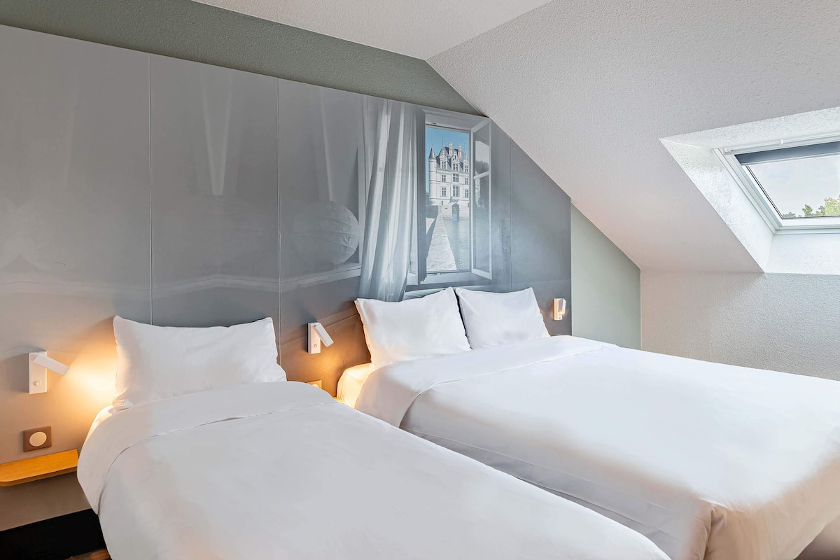 A hotel room with two beds, a large window with a view of a building, and a skylight.