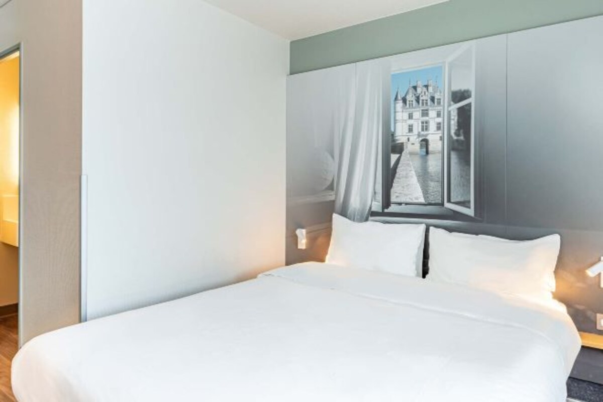 Una habitación de hotel con una cama, un ventanal con vista a un edificio y una mesita de noche con una lámpara.