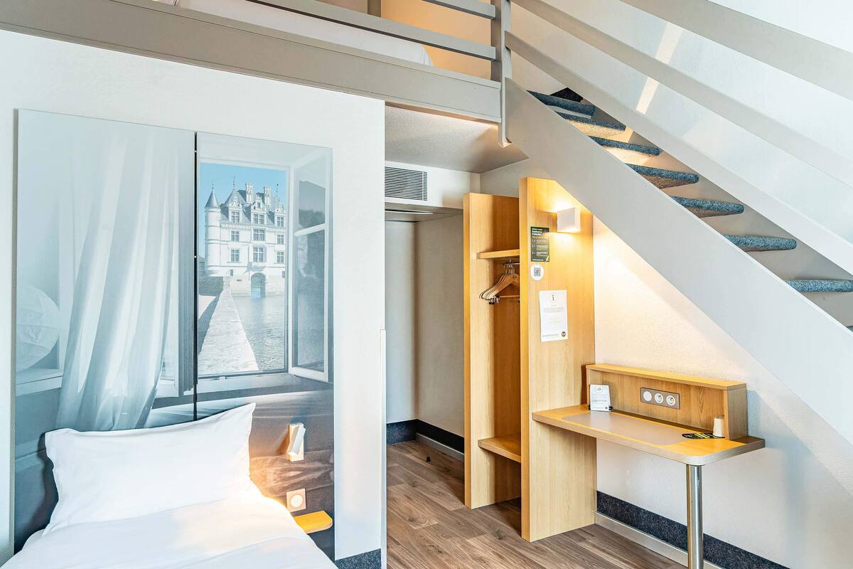 A hotel room with a bed, a wooden desk, a coat rack, and a staircase.