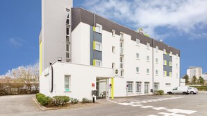 A multi-story hotel with a modern design, featuring a prominent entrance with a yellow pillar, a visible fire hydrant, and a parking area with a white car. - B&B HOTEL Tours Parc Expo St Avertin (Saint-Avertin)