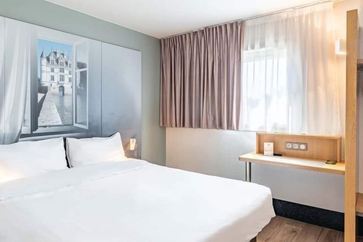 A hotel room with a large bed, a nightstand, a window with curtains, and a wall-mounted control panel.