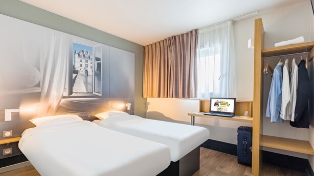 A hotel room with two beds, a desk with a laptop, a wardrobe, and a view of a building through a window. - B&B HOTEL Tours Parc Expo St Avertin (Saint-Avertin)