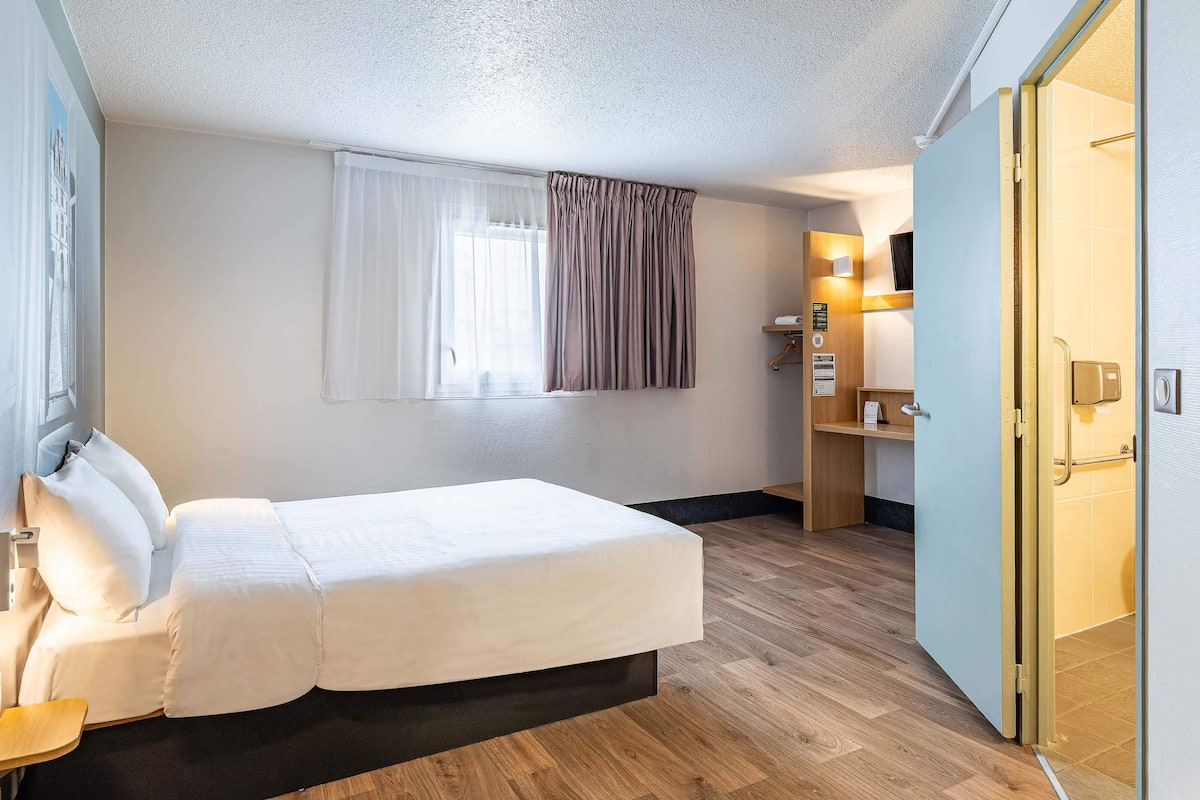 A hotel room with a bed, a window with curtains, a wooden floor, a bathroom with a shower, and a door.