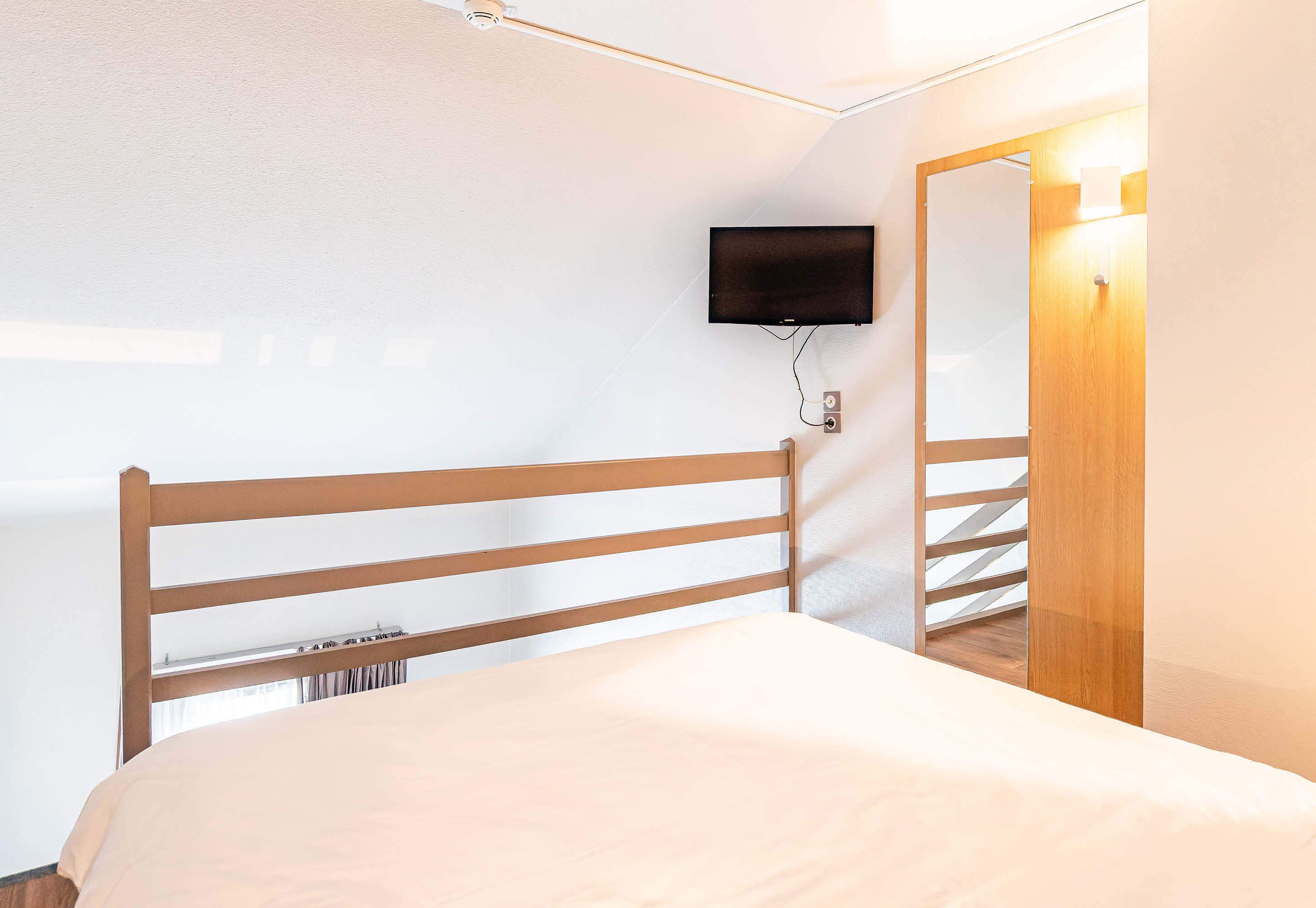 Un dormitorio con cabecera de madera, un televisor de pantalla plana montado en la pared y un espejo que refleja parte de la habitación.
