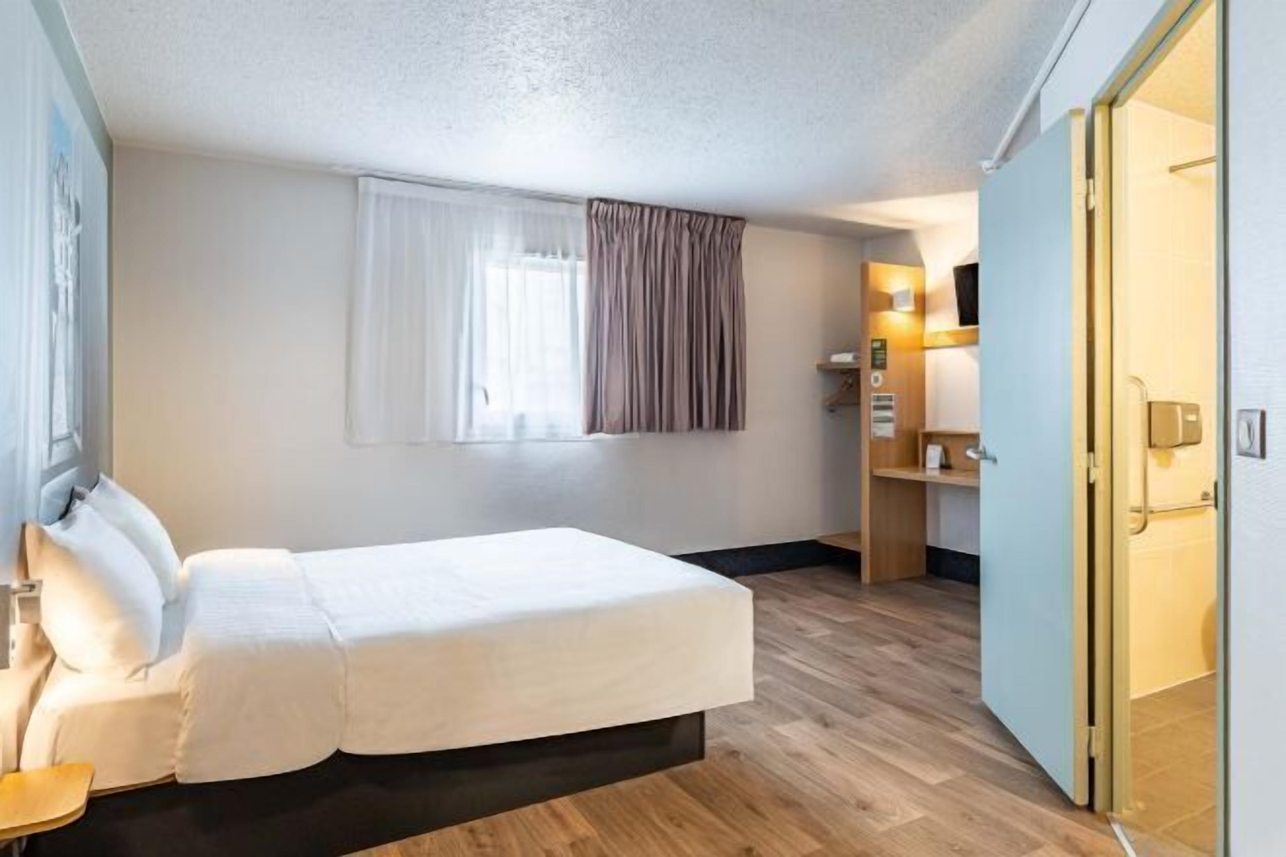 A hotel room with a bed, a window with curtains, a desk with a chair, and a door leading to the bathroom.