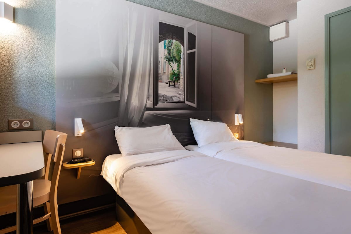 A hotel room with a bed, bedside table, chair, and a wall mural of a window with curtains.