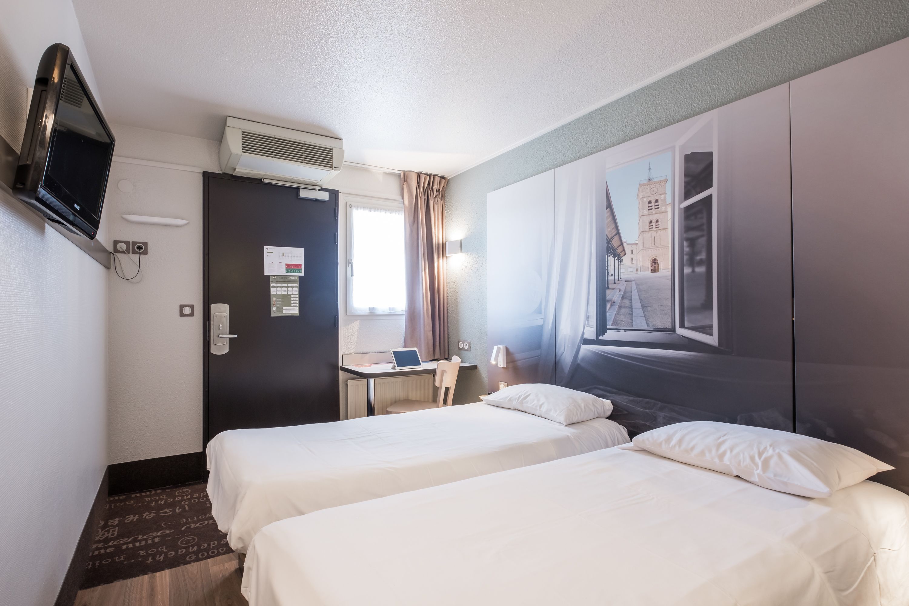 A hotel room with two beds, a desk with a laptop, a TV, and a large window with a view of a building.