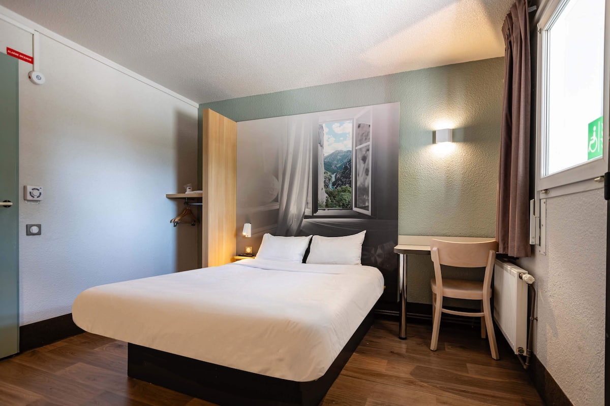 A hotel room with a bed, a small table, a chair, a window with a view of mountains, and a wall-mounted light.