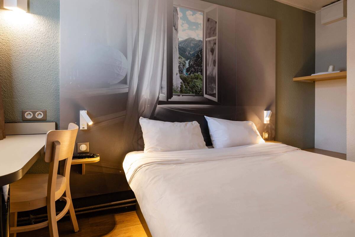 A modern hotel room with a large bed, a desk, and a chair. There is a window with a view of mountains and a river.