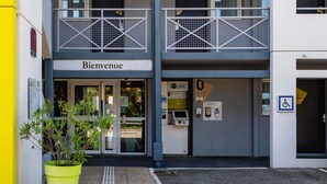 Een gevelingang met een 'Bienvenue'-bord, een toegankelijke ingang voor rolstoelen, en een bloempot in de voorgrond.