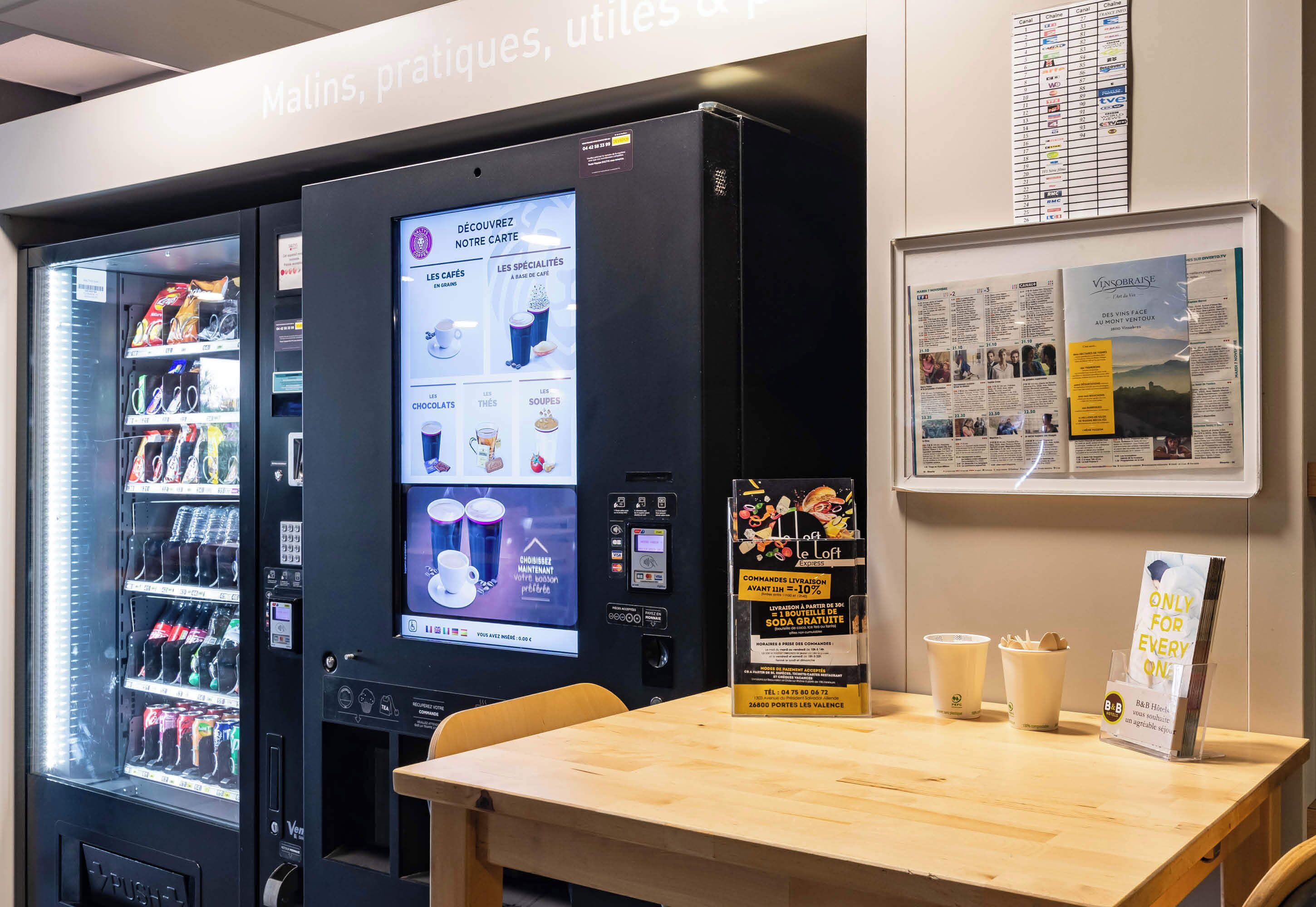 A self-service beverage machine with a digital menu, a seating area with a table, and informational posters on the wall.
