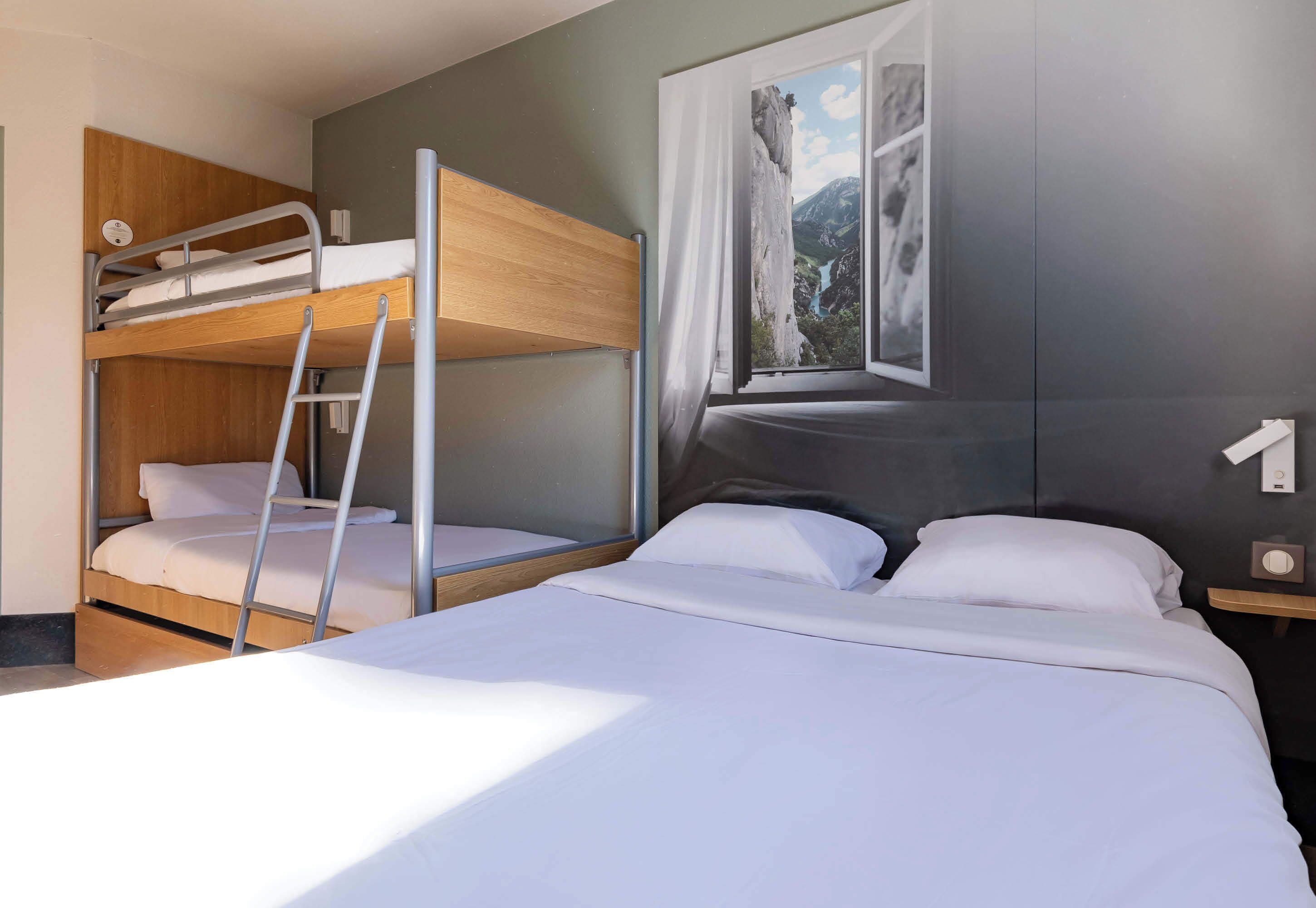 Habitación de hotel con literas, una cama individual, ventana con vistas a la montaña y mesita de noche con lámpara.